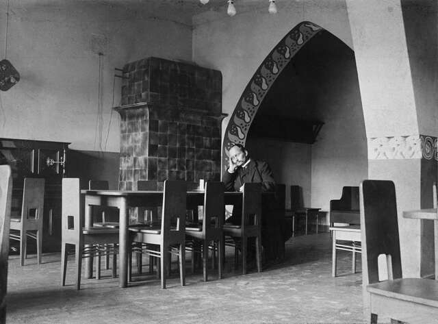 Black and white picture of a man reading in an room with many tables and chairs.