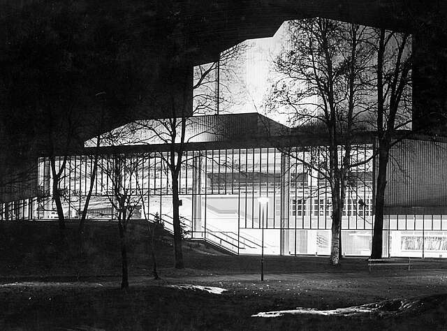 Black and white image of the facade with its glass walls illuminated during the evening.