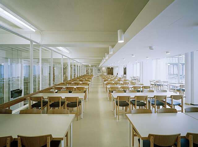 White room with glass walls. White tables are surrounded by wooden chairs.