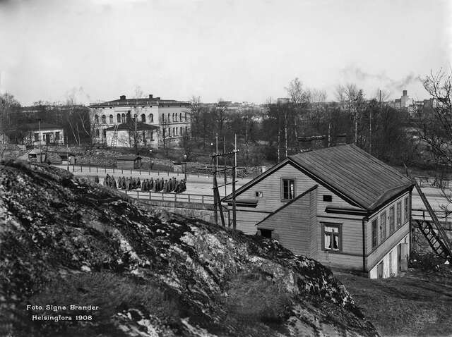 Black and white picture of the villa viewed from a rock. On the street there are rows of soldiers marching in long coats.