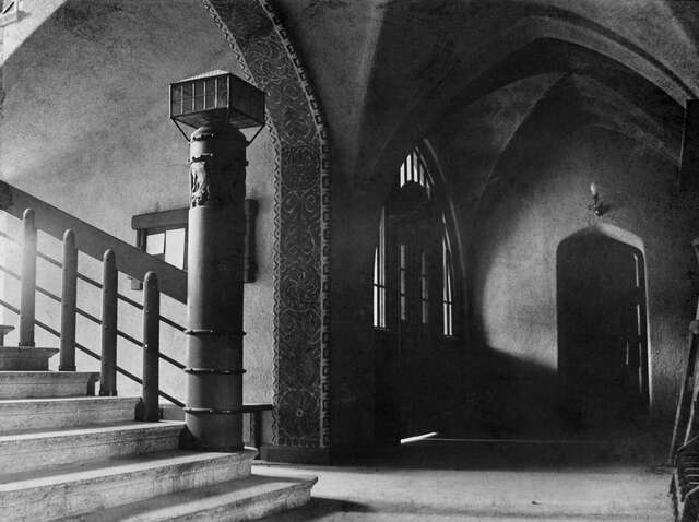 Black and white picture of a dark hallway with a groin vault ceiling and paintings in an arch above the stairs.