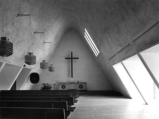 Bare altar wall with a hanging cross, bench rows and hanging light fixtures on the left.