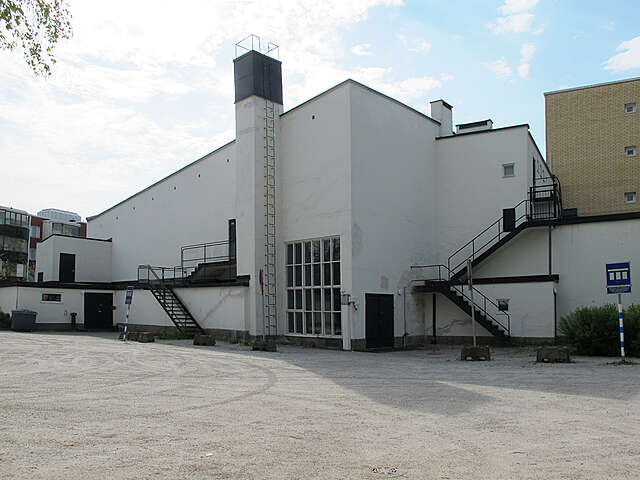 The back of the white building with a fire escape, a small window wall and parking space behind the building.