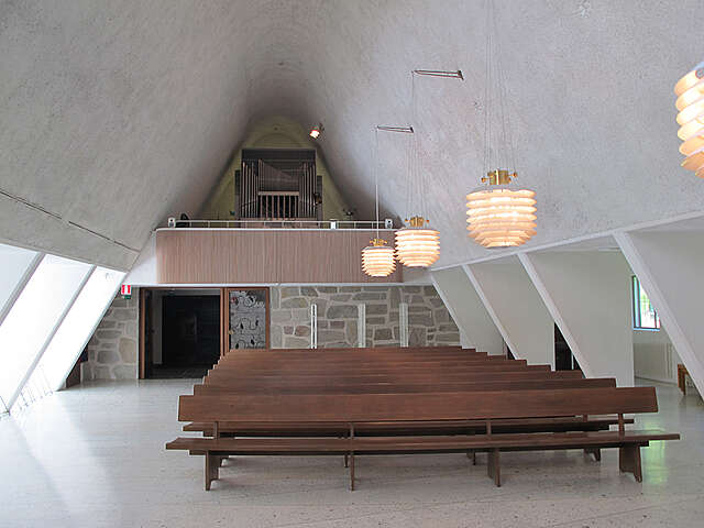 View away from the altar towards the back. The organ is on a balcony, benches are on the left with lamps hanging above them from the curved white plastered ceiling.