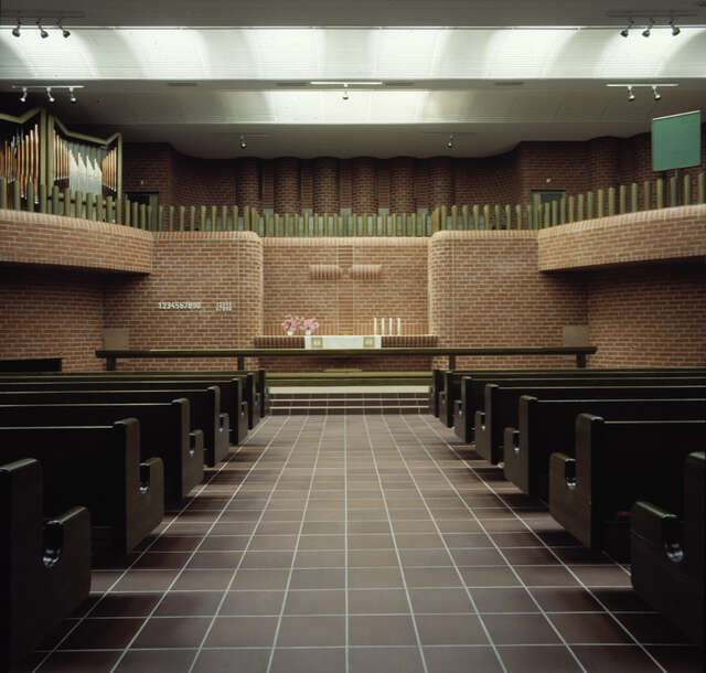 Dark church interior with red. brick walls, dark brown tile floors and black benches.