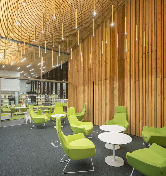 Green armchairs around tables next to a wooden panel wall and a hanging light installation above them.