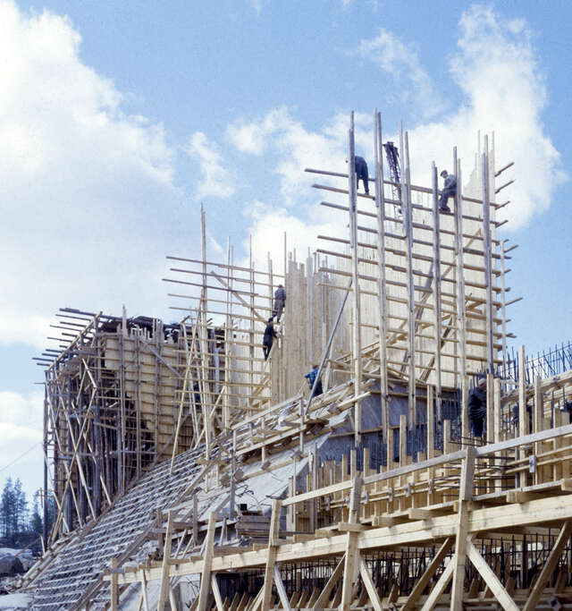Photo taken from the construction site, showing three construction site workers climbing high wooden scaffolding.