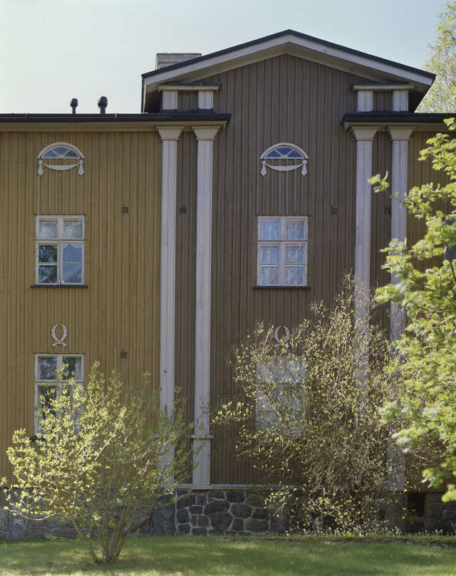 Two-storey wooden house with decorative details, bushes in front of it.