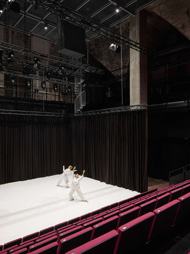 Theater stage with white floor. Two people dressed in white stand on stage. View to the stage is from the stands.