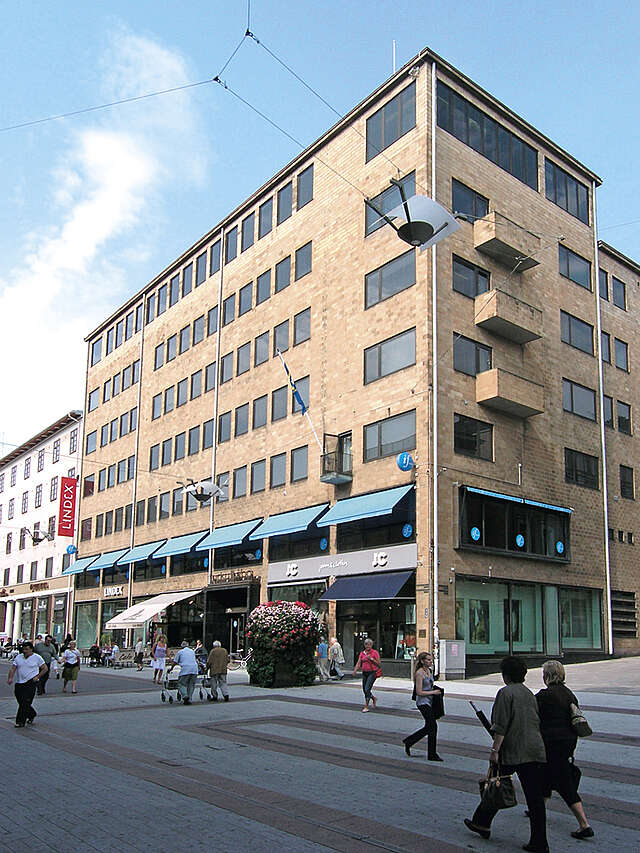 Brick building with square windows, stores on the first floor, people walking along the trees tin from of it.