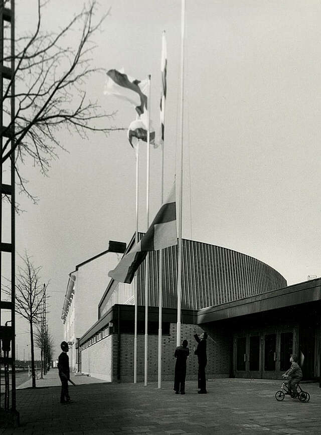 Min entrance with for flagpoles in front of it. Two men are raising the Finnish flag up the pole.