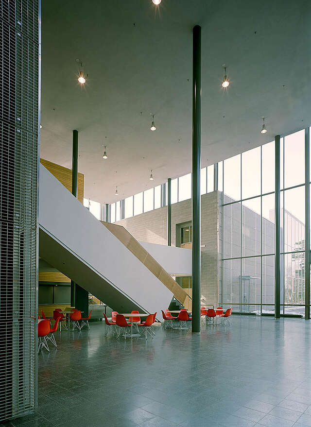 Spacious lobby with glazed walls, grey support columns and round tables for the students to sit at.