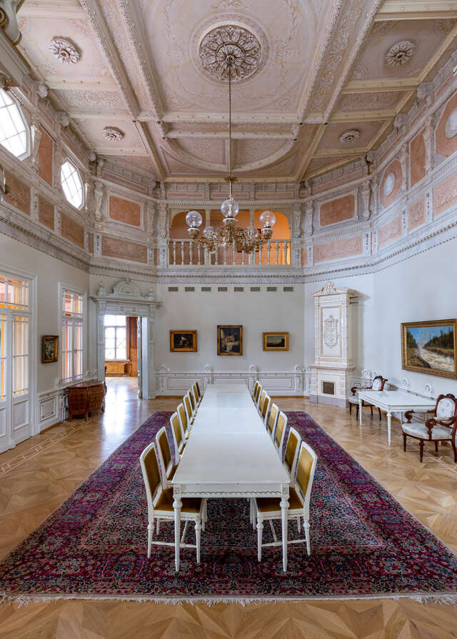 Old, decorative space with light-coloured walls and ceiling, wood parquet floor and a very long table with chairs in the middle