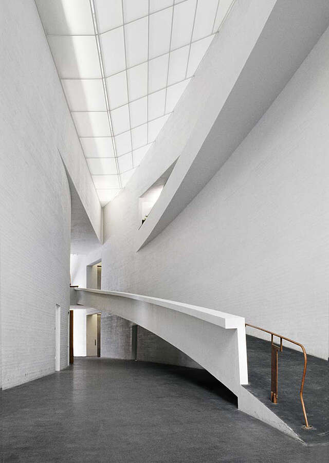Spaceious entrance lobby with white tile walls, grey stone floors and white glass ceiling letting in some light. At the right there is a long ramp leading to the second floor.