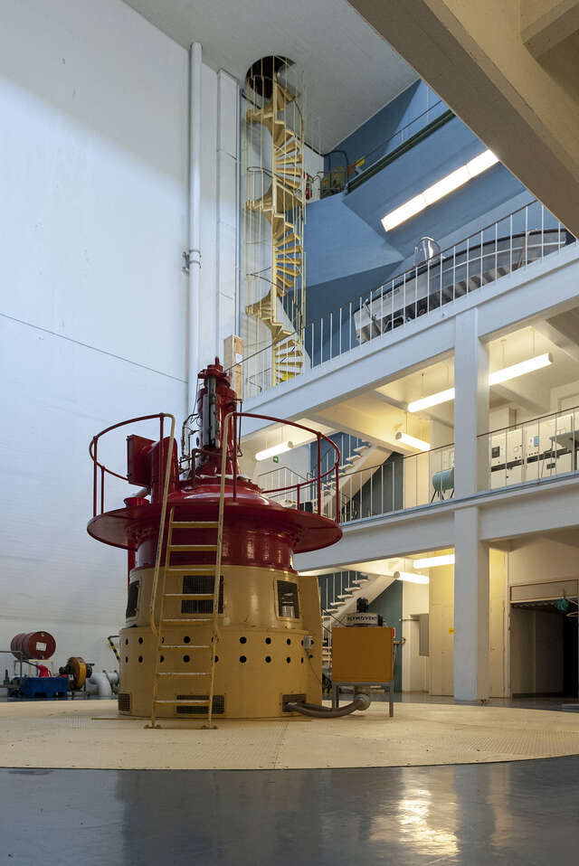 Picture from the machine hall with machinery decorated with bright red and bright yellow colours. In the background there is a bright yellow spiral staircase.