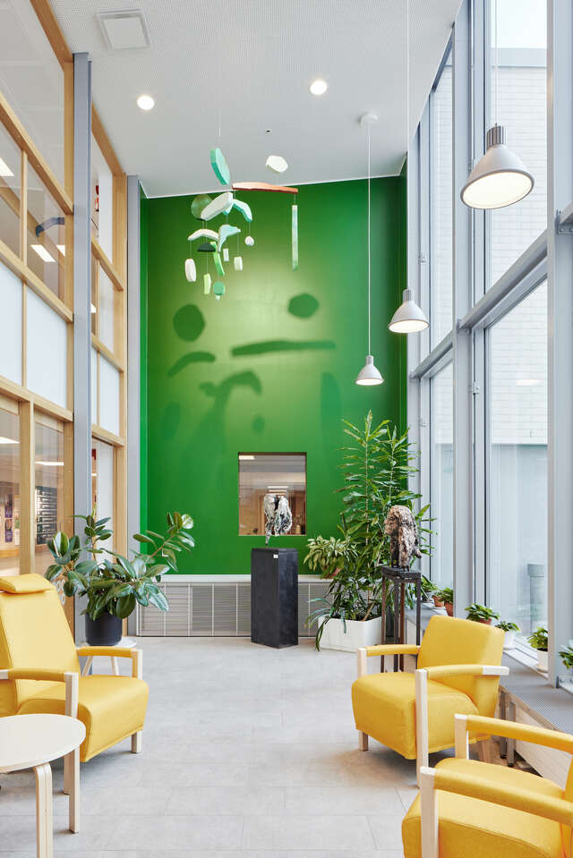 The lobby of a large building, with a green wall, large glass windows and yellow chairs