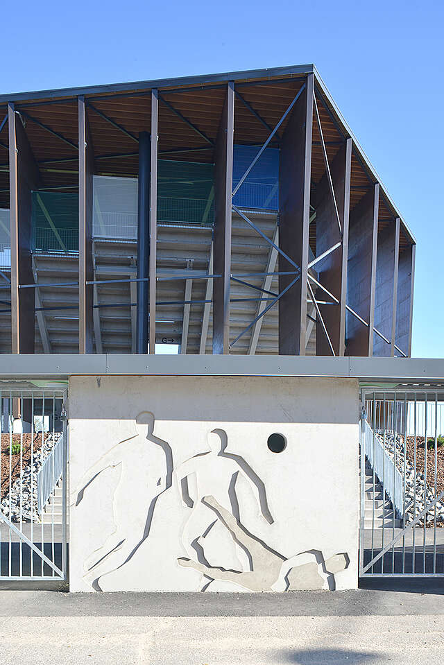 Side of the spectator stand with a fence and a sculpture of football players.