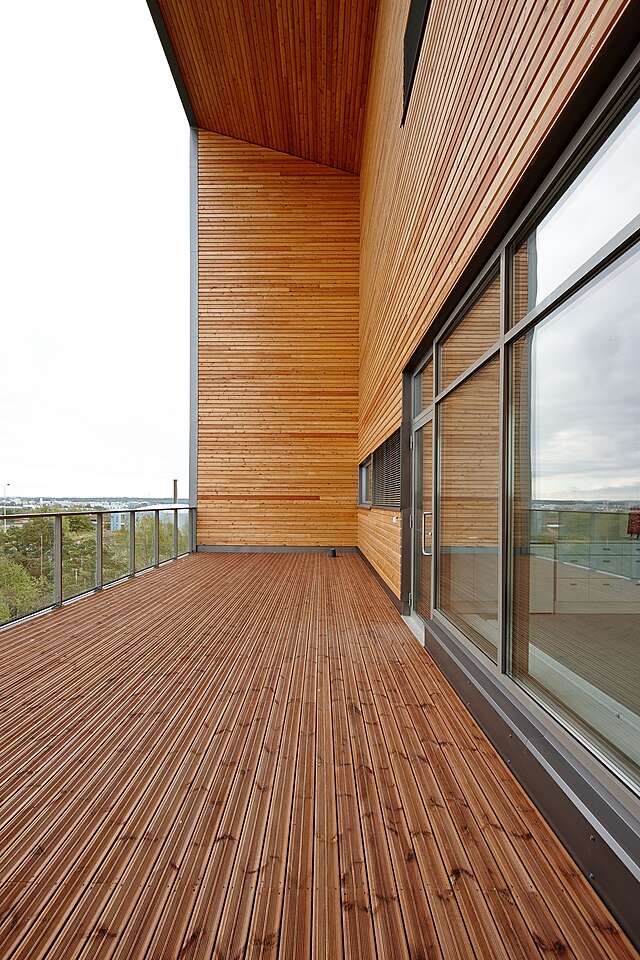 Large balcony with a glass railing, warm wooden floor and sides and large glass windows to the inside.