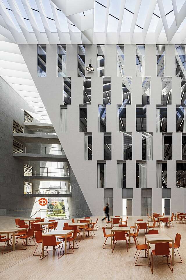 Hall with tables and chairs, a white wall with long glass windows and a view to different story hallways.