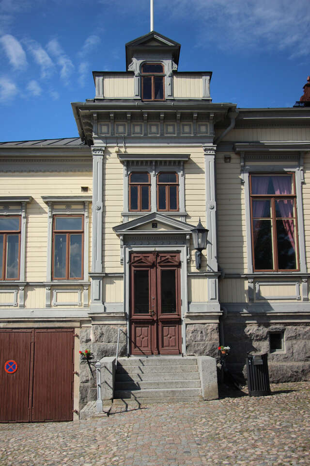 Entrance to the building. The building has a stone base, yellow wooden boards on the facade and grey decoraional elements around the corners door and windows.