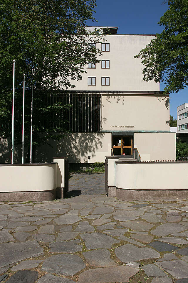 Entrance to theTurku library with cream colored fences, three flagpoles and trees in front of the cream colored library building.