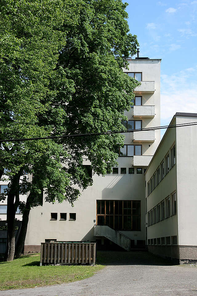 White facade with several small windows and balconies next to a large tree.