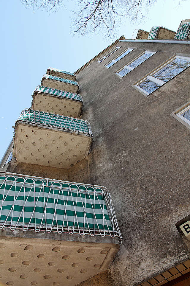 Multiple story building with a grey plaster facade and balconies with white metal railings covered from the inside with a green and white striped fabric.