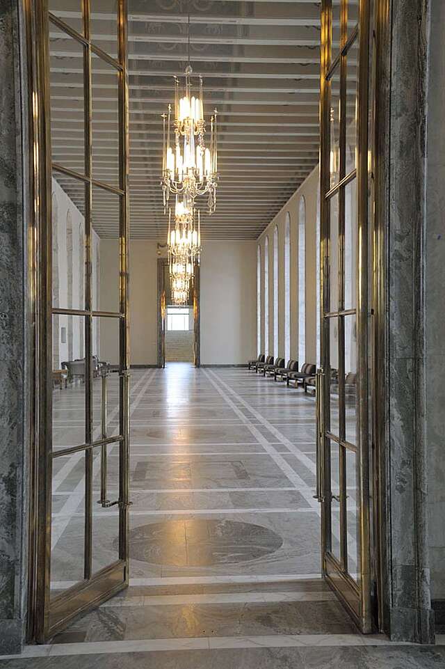 View through glass doors into a long room with marble floors an golden chandeliers.