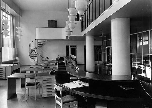 Mustavalkoinen kuva, jossa näkyy pöytiä ja portaat yläkertaan.