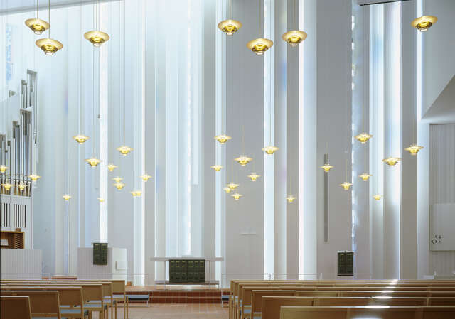 Pakila Church of the Good Shepherd Church. Minimalistic and high organ are on the left. Instead of an altarpiece, behind the altar is a white wall with various recesses from which natural light flows in. The church hall is decorated with golden clusters of lamps hung at different heights.